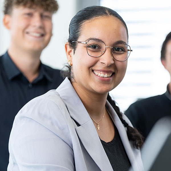 The person is wearing a grey blazer and has curly hair. In the background, there are others dressed in dark clothing.