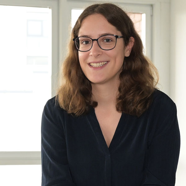 A person with curly brown hair sits in a bright room with windows in the background.