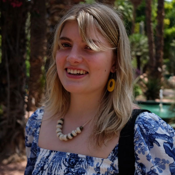 A person with wavy blonde hair wears a floral patterned top and a beaded necklace, surrounded by tropical plants.