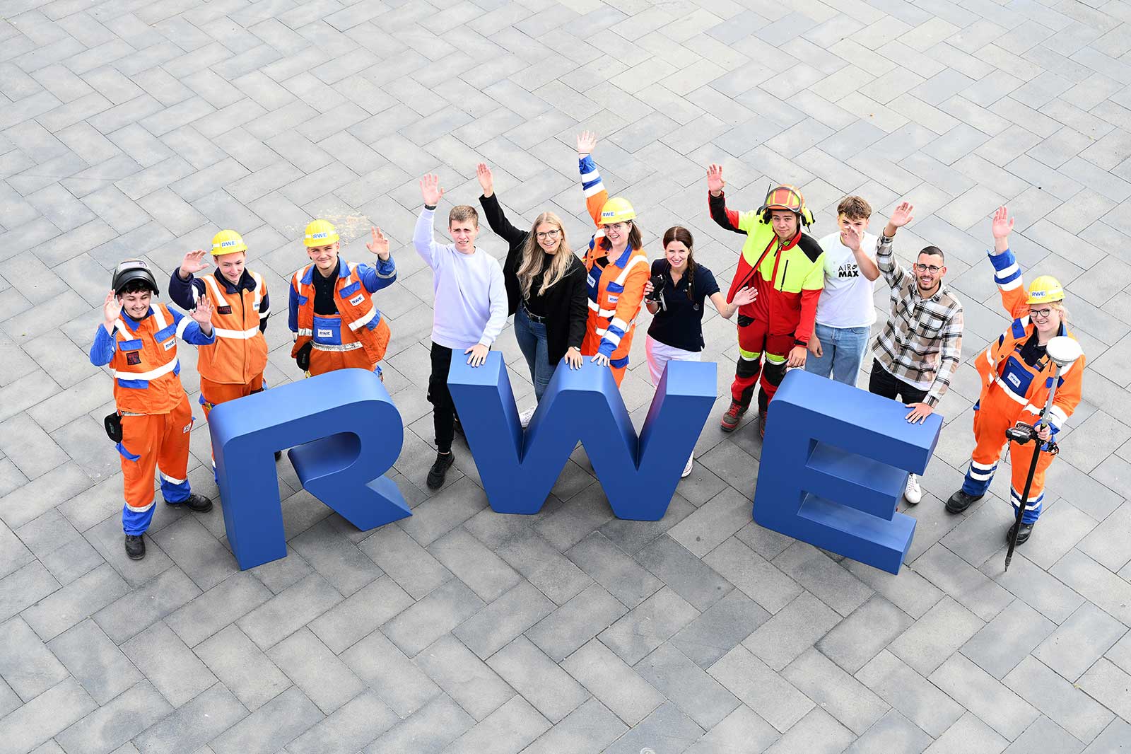 Eine vielfältige Gruppe von Arbeitern in Sicherheitskleidung posiert fröhlich um große blaue Buchstaben, die 'RWE' buchstabieren.
