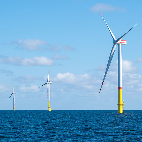 Im Vordergrund steht ein hoher weißer Offshore-Windturbinenmast aus Stahl mit gelber Basis und roten Blattspitzen, umgeben von blauem Meer und klarem Himmel.
