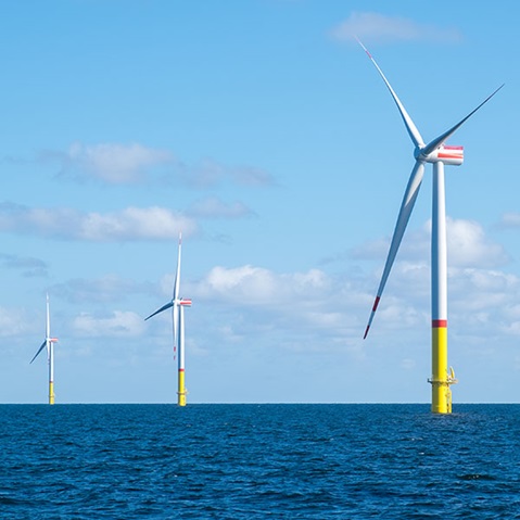 Im Vordergrund steht ein hoher weißer Offshore-Windturbinenmast aus Stahl mit gelber Basis und roten Blattspitzen, umgeben von blauem Meer und klarem Himmel.