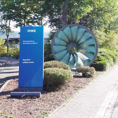 Ein Willkommensschild von RWE steht neben einer großen Turbinen-Skulptur in einem schönen Landschaftsbereich.