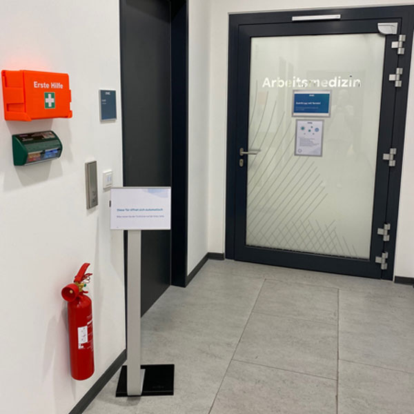 Ein Eingangsbereich einer Arztpraxis mit einem Erste-Hilfe-Kasten, Feuerlöscher und einem Schild mit dem Büro-namen 'Arbeitsmedizin'.