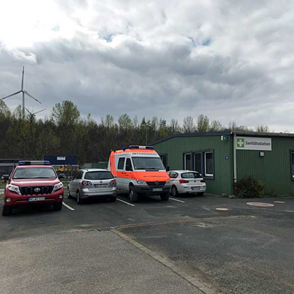 Ein grünes Sanitärgebäudes mit verschiedenen Fahrzeugen, darunter ein Krankenwagen, unter einem bewölkten Himmel geparkt.
