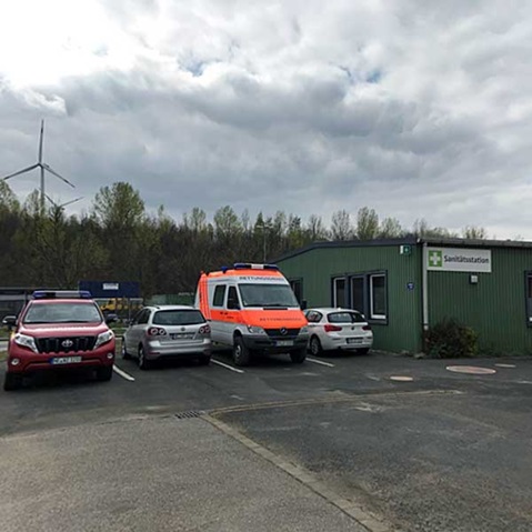 Ein grünes Sanitärgebäudes mit verschiedenen Fahrzeugen, darunter ein Krankenwagen, unter einem bewölkten Himmel geparkt.
