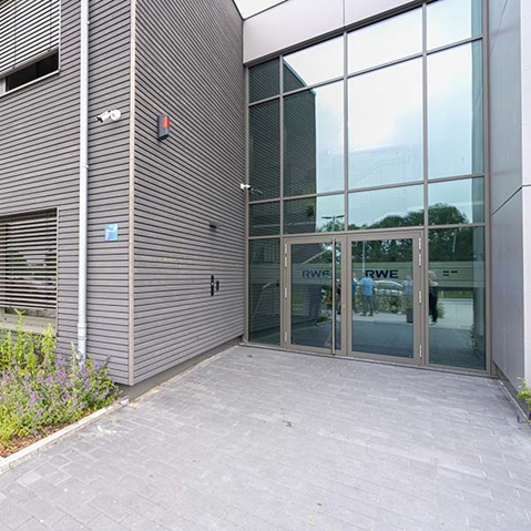 Eingangsbereich eines modernen Bürogebäudes mit grauer Metallfassade und großen Glastüren, auf denen „RWE“ zu sehen ist, mit einem Blumenbeet links.