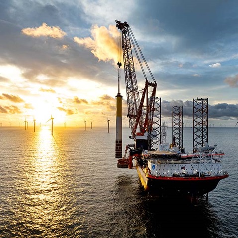 Ein großes Installationsschiff mit weiß-rotem Rumpf und hohen Stahlkranen liegt im Vordergrund vor einem Offshore-Windpark, goldenes Sonnenlicht spiegelt sich auf ruhigem Meer und bewölktem Himmel.