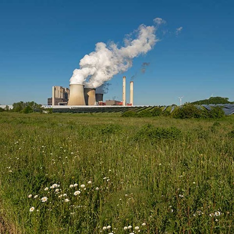 Ein Kraftwerk, das Dampf unter einem klaren blauen Himmel ausstößt, umgeben von grünen Feldern und Solarpanelen.