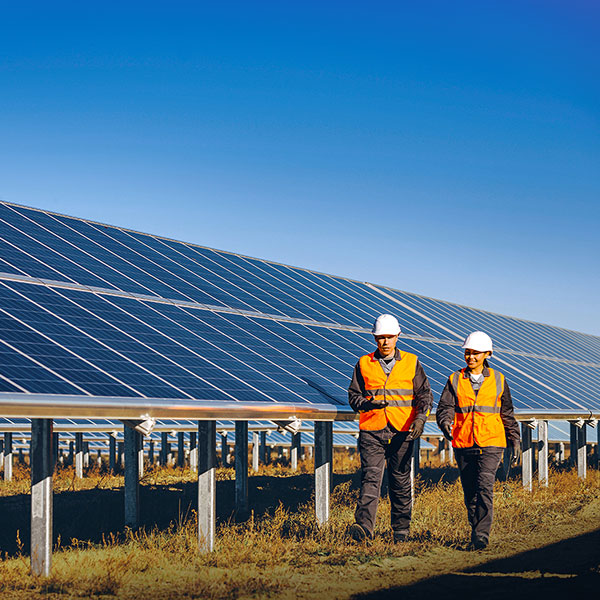 Zwei Arbeiter in Sicherheitskleidung gehen zwischen Solarpanels unter einem klaren blauen Himmel.