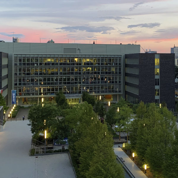 Ein modernes Gebäude mit großen Glasfenstern, umgeben von Grünflächen und beleuchteten Wegen, bei Sonnenuntergang.
