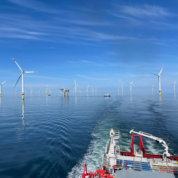 Vom Heck eines Schiffes mit rotem Geländer und weißer Ausrüstung führt eine schimmernde Fahrspur über das ruhige, spiegelglatte blaue Meer zu weißen Windturbinen.