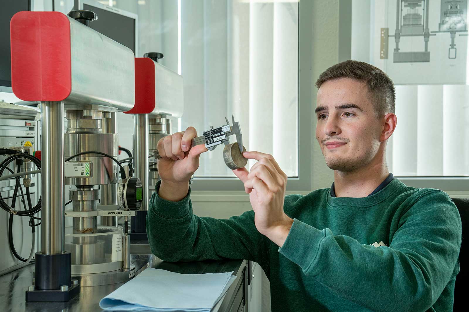 Eine Person in einem grünen Pullover misst ein rundes Objekt mit einer Messschieber in einem Labor.