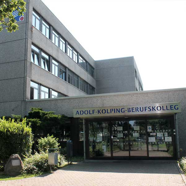 Der Eingang des Adolf-Kolping-Berufskollegs, mit einem modernen Gebäude, grüner Bepflanzung und klarem blauen Himmel.