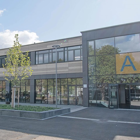 Ein modernes Gebäude mit großen Fenstern, einem Baum im Vordergrund und einem auffälligen Buchstaben A an der Wand.