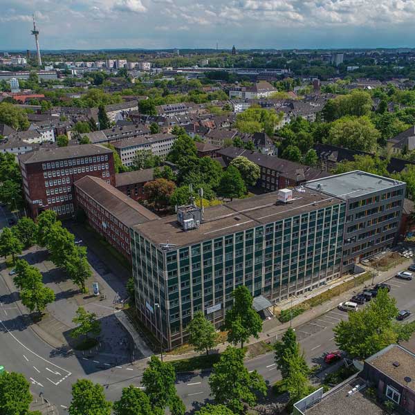 Luftaufnahme eines modernen Gebäudes mit Glasfassade, umgeben von Bäumen und Wohngebieten unter einem bewölkten Himmel.