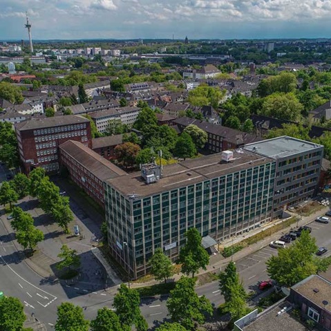 Luftaufnahme eines modernen Gebäudes mit Glasfassade, umgeben von Bäumen und Wohngebieten unter einem bewölkten Himmel.