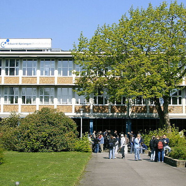 Eine Gruppe von Menschen versammelt sich vor einem modernen Backsteingebäude mit einem Baum und Sträuchern im Vordergrund unter klarem blauen Himmel.