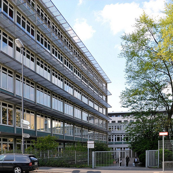 Ein modernes Gebäude mit vielen Glasfenstern, umgeben von Grünanlagen und Wegen, unter einem klaren blauen Himmel.