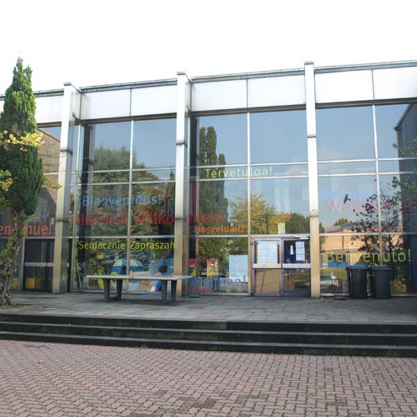 Ein moderner Eingang eines Gebäudes mit Glasfassade und bunten Begrüßungsschildern in mehreren Sprachen.