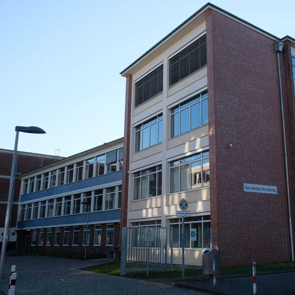 Ein modernes Bildungsgebäude mit großen Fenstern und einer Backsteinfassade, das einen eingezäunten Eingang und Lichtmasten aufweist.