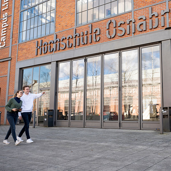Studierende gehen vor dem Gebäude der Hochschule Osnabrück, das große Glastüren und eine moderne Backsteinfassade aufweist.