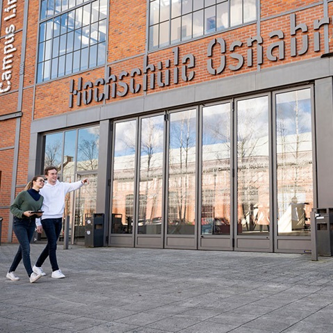 Studierende gehen vor dem Gebäude der Hochschule Osnabrück, das große Glastüren und eine moderne Backsteinfassade aufweist.
