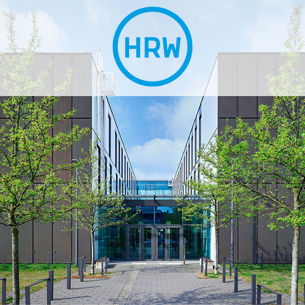 Ein moderner Gebäudeeingang, flankiert von jungen Bäumen, unter einem klaren blauen Himmel.