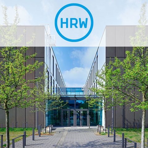 Ein moderner Gebäudeeingang, flankiert von jungen Bäumen, unter einem klaren blauen Himmel.