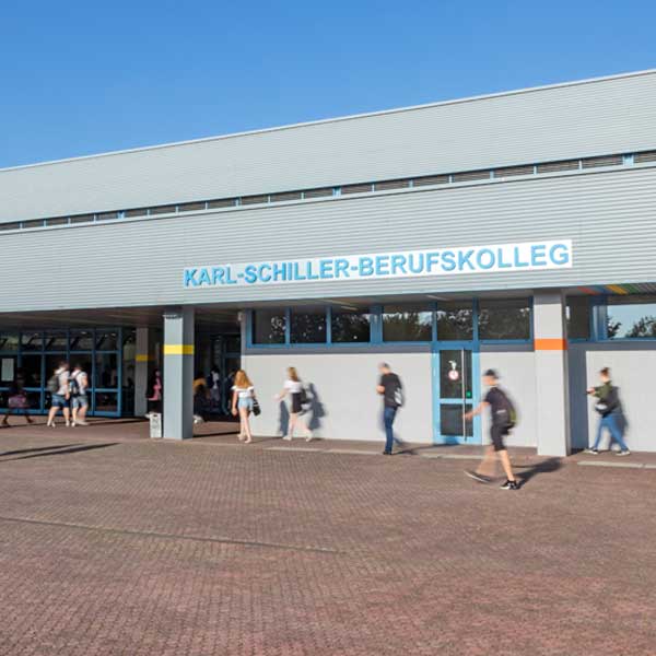 Die Fassade des Karl-Schiller-Berufskollegs mit Schülern, die vorbeigehen, und klarem blauen Himmel.