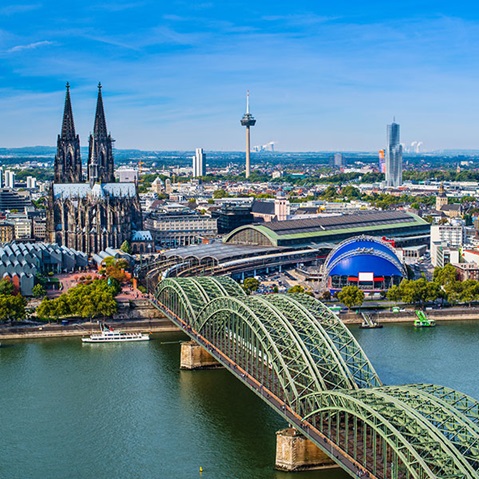 Ein Panoramablick auf Köln mit dem Dom, einer Brücke und dem Rhein unter einem klaren blauen Himmel.