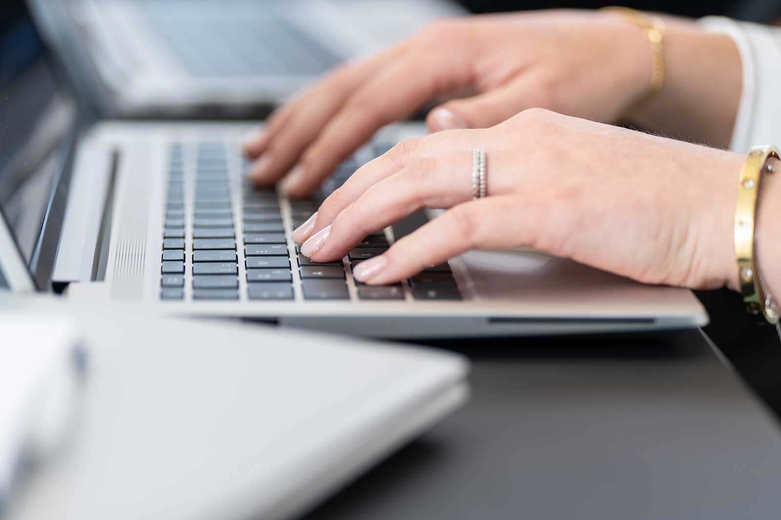 Eine Nahaufnahme von Händen, die auf einer Laptop-Tastatur tippen. Eine Hand trägt einen silbernen Ring.