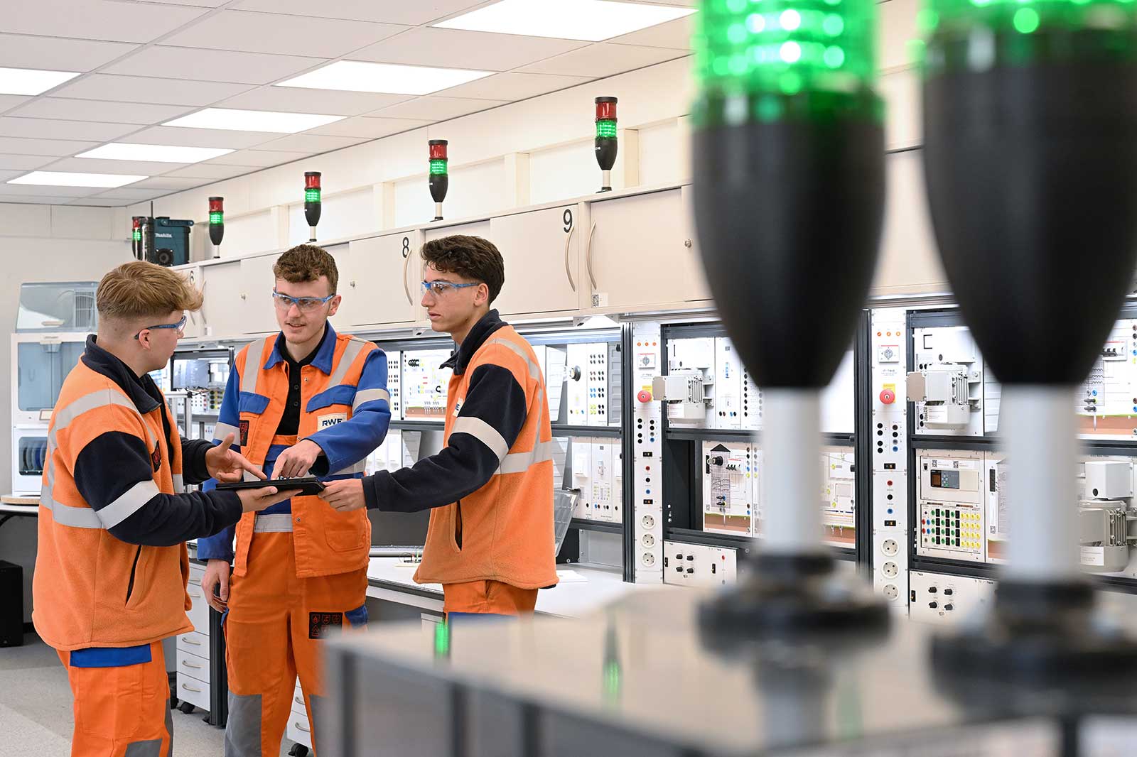 Drei Techniker in orangefarbenen Uniformen besprechen sich über ein Tablet in einem hochmodernen Kontrollraum voller Geräte.