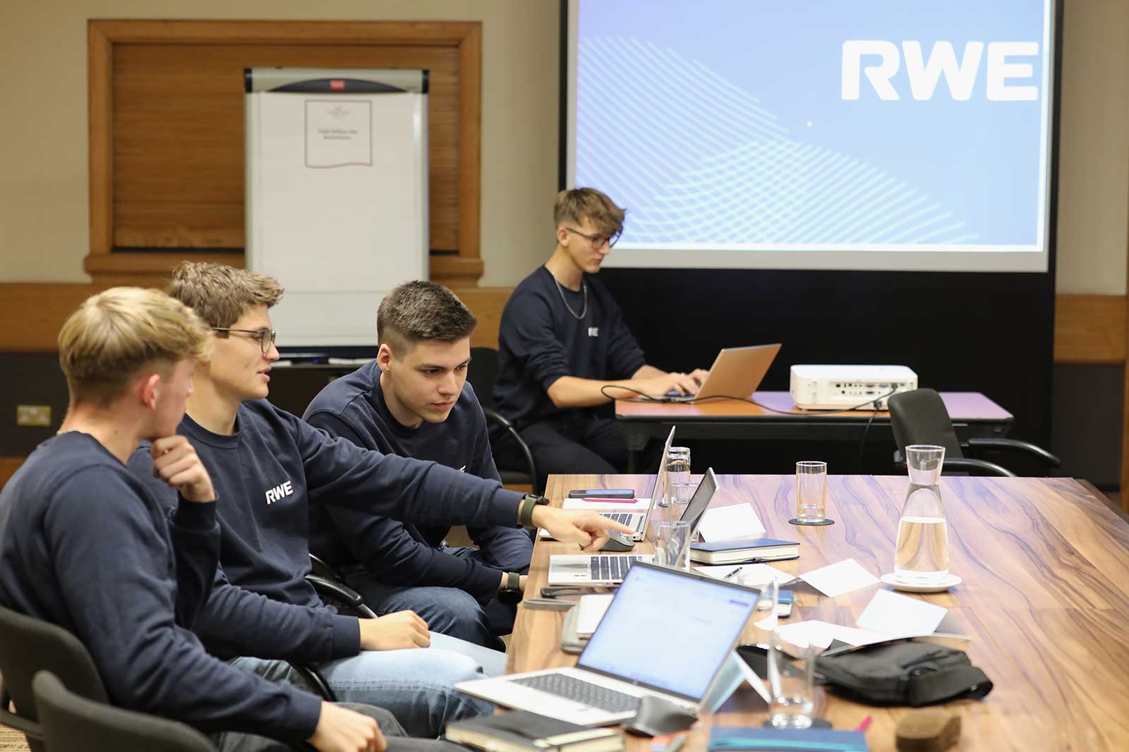 Eine Gruppe von Personen in einem Besprechungsraum, die an Laptops zusammenarbeiten. Ein Projektor zeigt das RWE-Logo an der Wand.