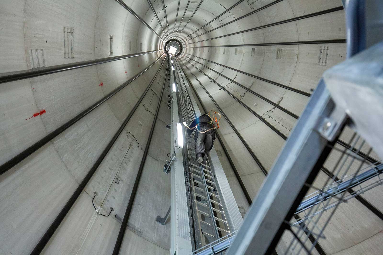 Ein Arbeiter klettert eine Leiter in einem großen, zylindrischen Gebäude mit Betonwänden und metallischen Stützen hinauf.