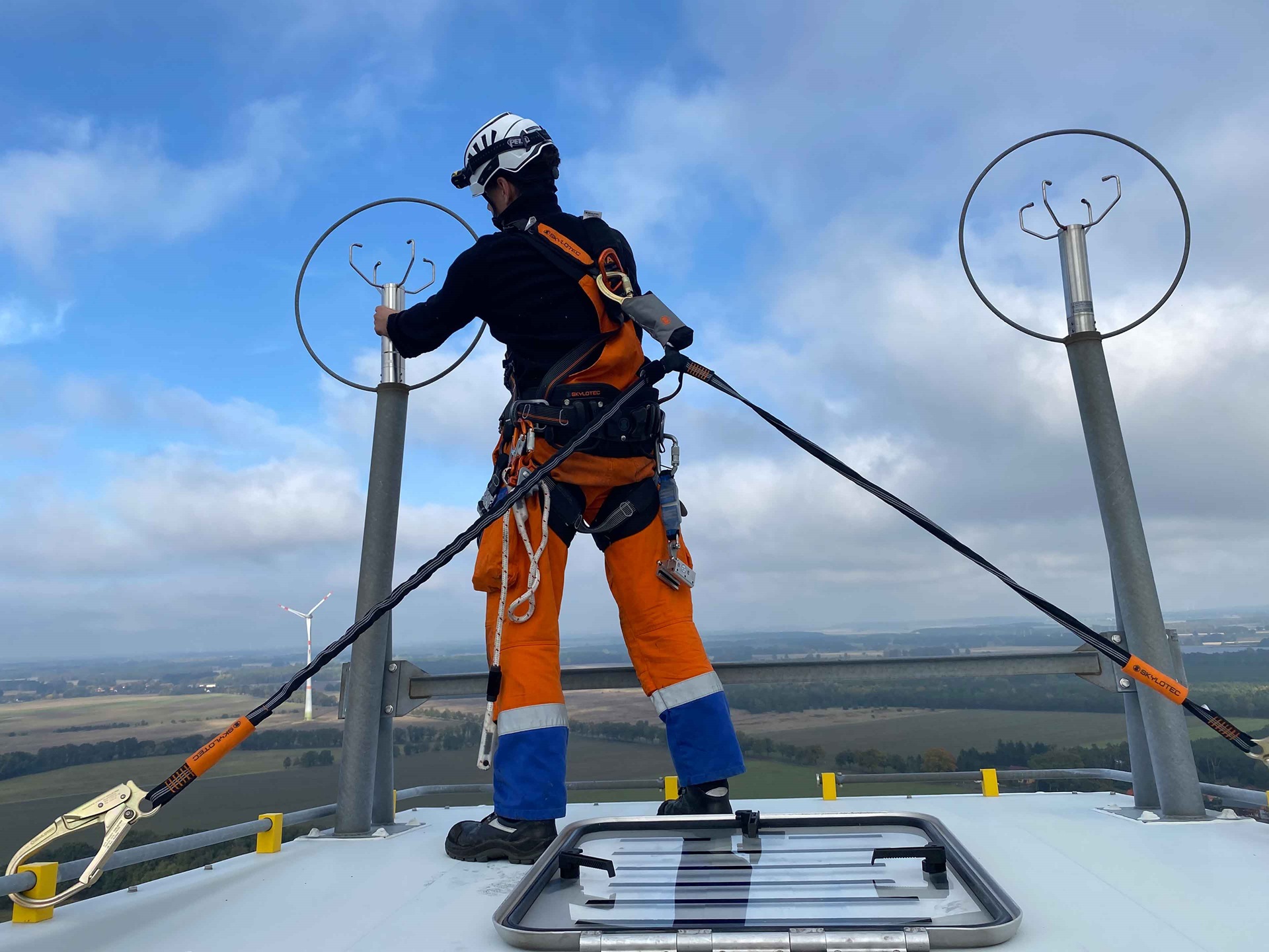 Ein Techniker in orangefarbener Uniform arbeitet an Geräten auf einem Windrad, mit einer weiten Landschaft darunter.