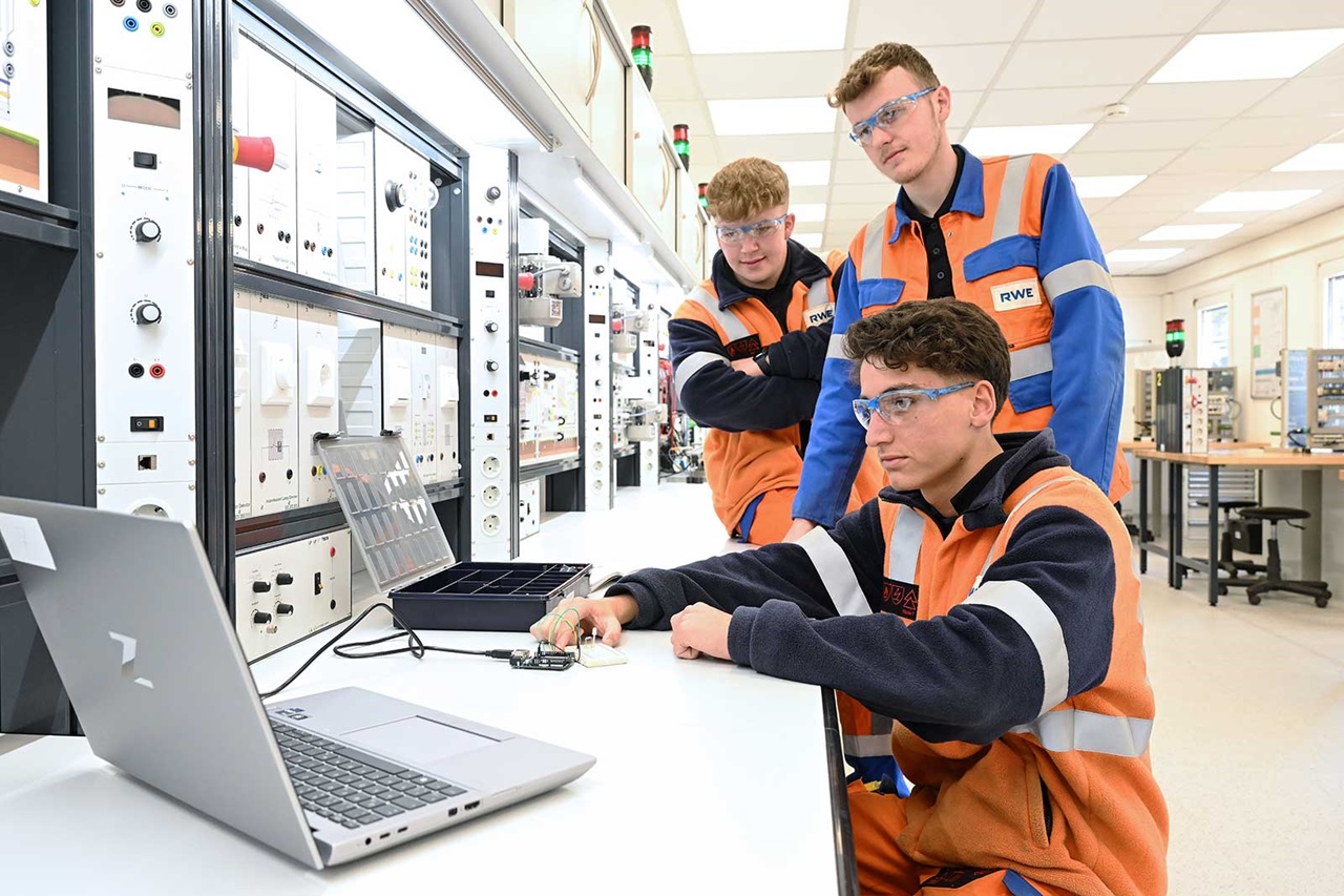 Drei Techniker in Sicherheitswesten arbeiten an einem Labortisch mit einem Laptop und Gerätschaften in einer modernen Einrichtung.