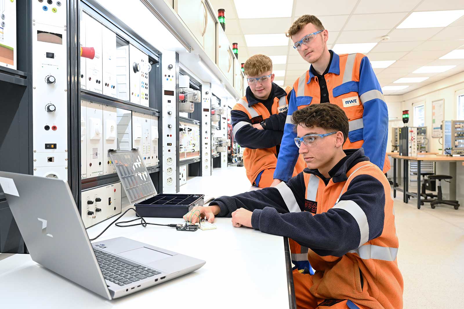 Drei Techniker in Sicherheitswesten arbeiten an einem Labortisch mit einem Laptop und Gerätschaften in einer modernen Einrichtung.