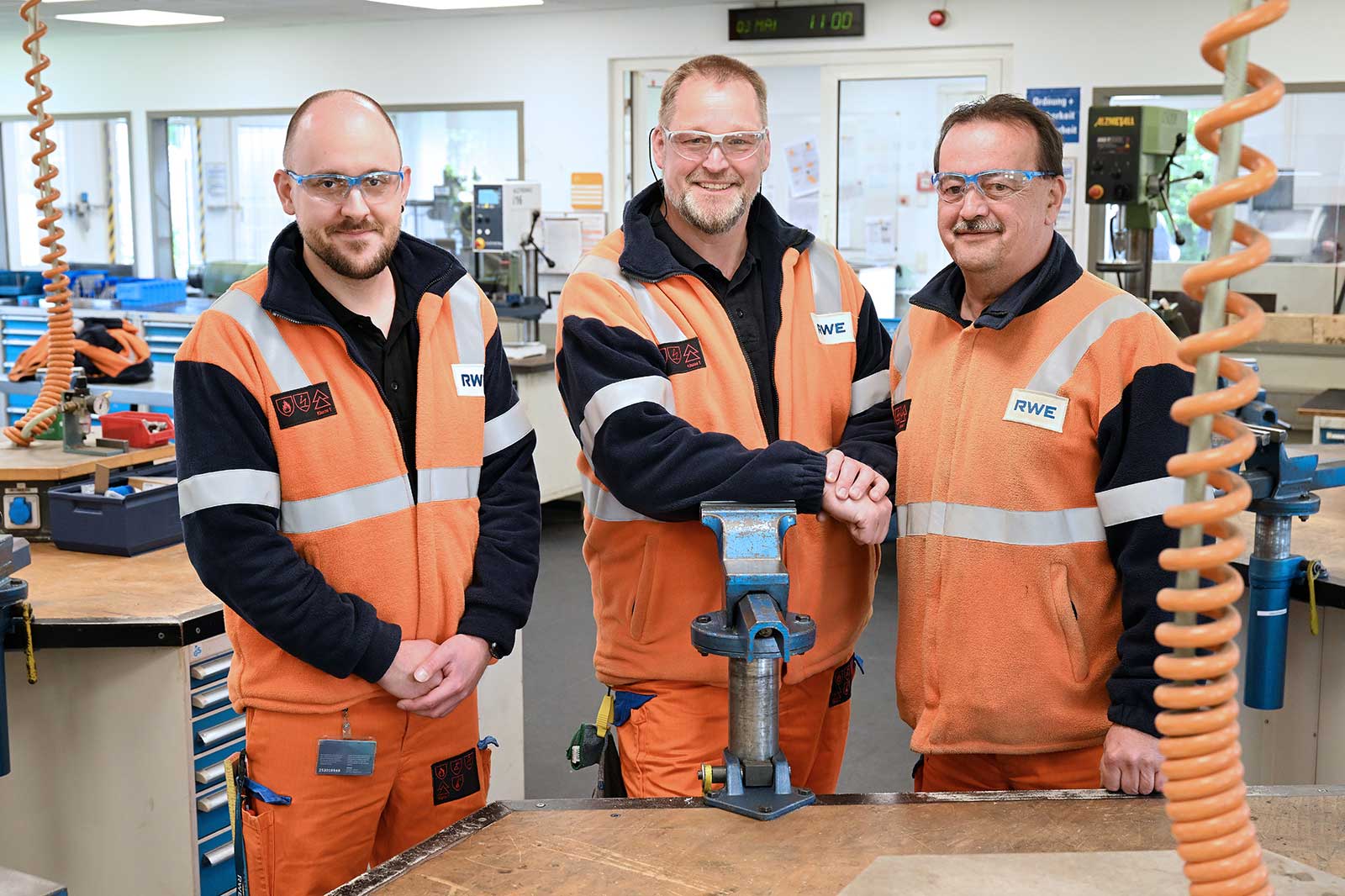 Drei Arbeiter in orangefarbenen und navy Jacken schütteln sich die Hände in einer Werkstatt mit Werkzeugen und Equipment im Hintergrund.