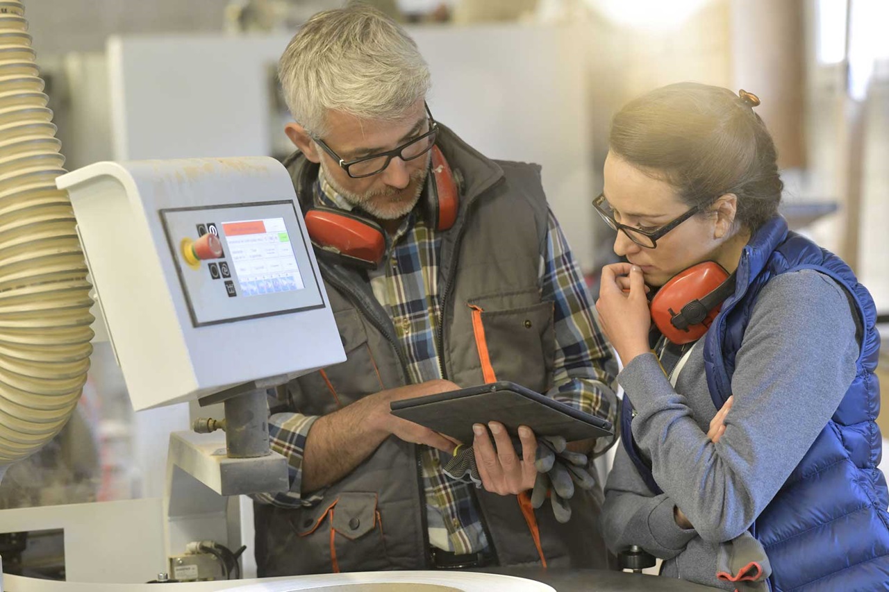 Zwei Arbeiter besprechen die Einstellungen einer Maschine, während sie Daten auf einem Tablet in einer Werkstatt analysieren.