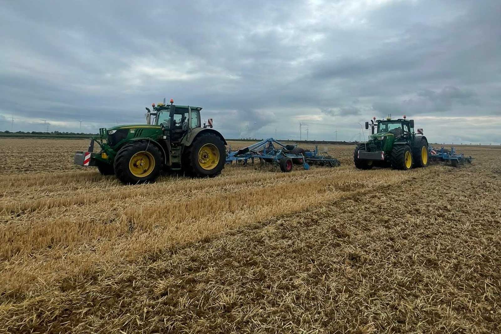 Zwei grüne Traktoren arbeiten in einem geernteten Feld unter einem bewölkten Himmel.