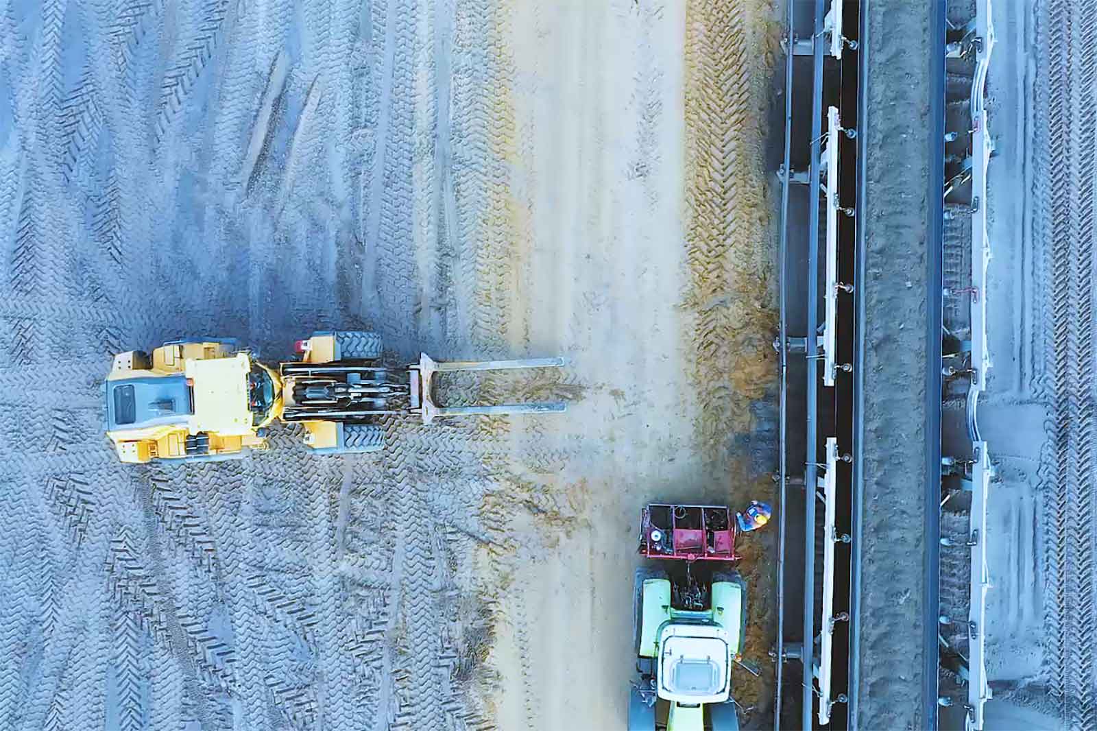 Vogelperspektive auf Baumaschinen auf einer sandigen Fläche mit Reifenspuren und einem grünen Traktor neben einem gelben Lader.
