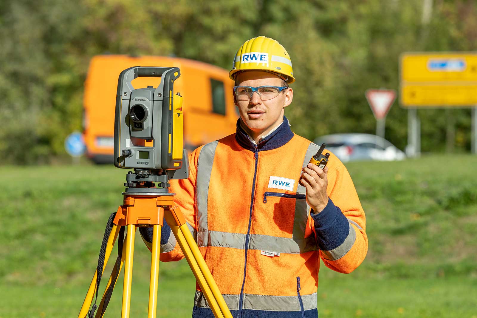 Ein Vermessungsfachmann trägt eine gut sichtbare Jacke und einen Helm und verwendet ein Totalstation auf einer Grasfläche.