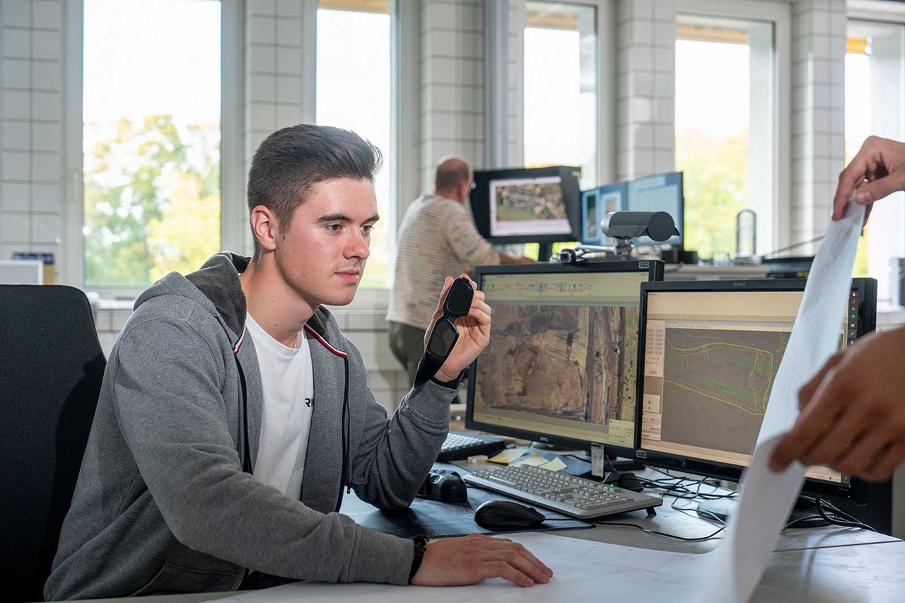 Eine Person sitzt an einem Schreibtisch und hält ein Gerät, während mehrere Computerbildschirme Karten und Grafiken in einem hellen Büro anzeigen.