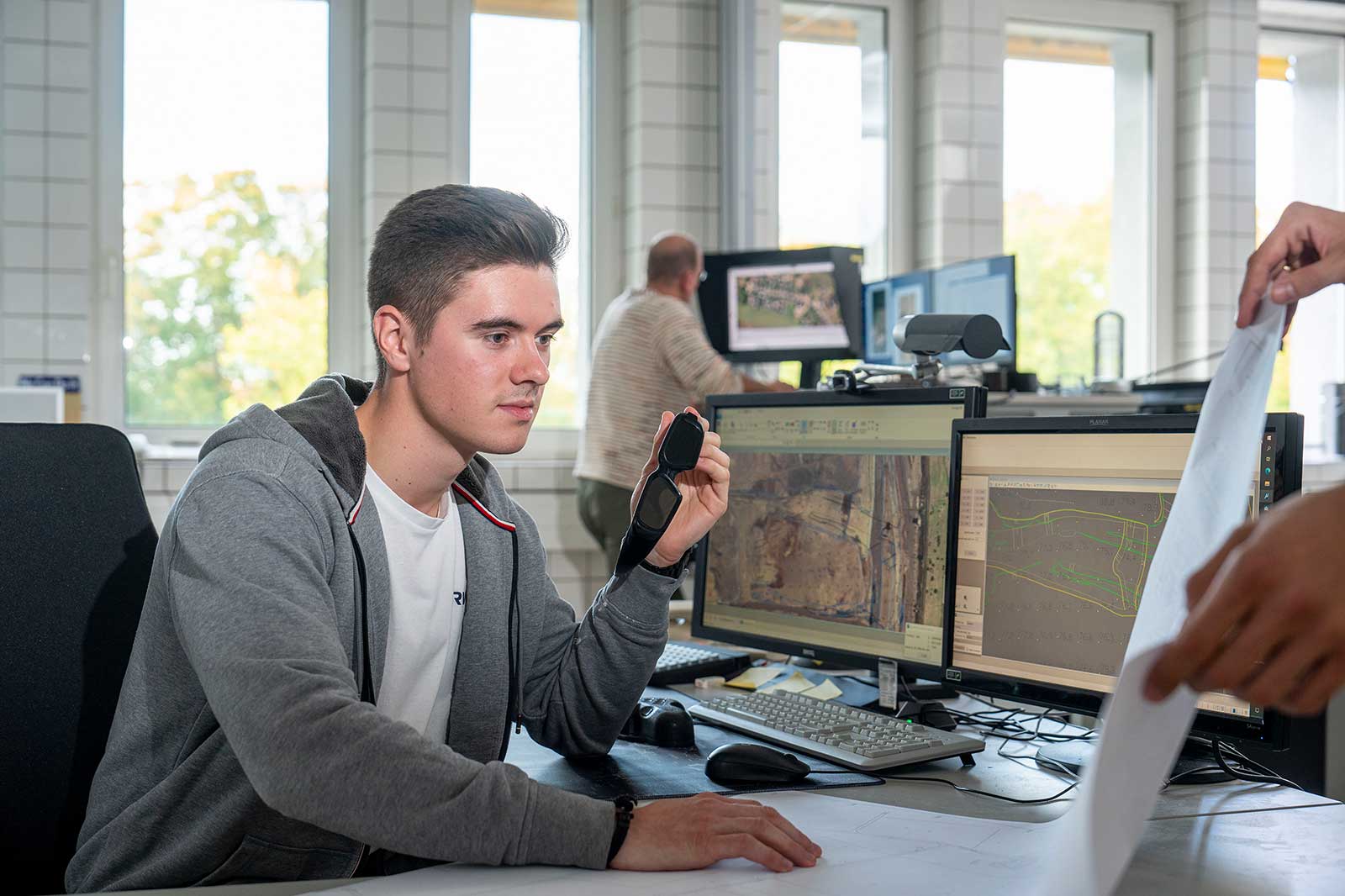 Eine Person sitzt an einem Schreibtisch und hält ein Gerät, während mehrere Computerbildschirme Karten und Grafiken in einem hellen Büro anzeigen.