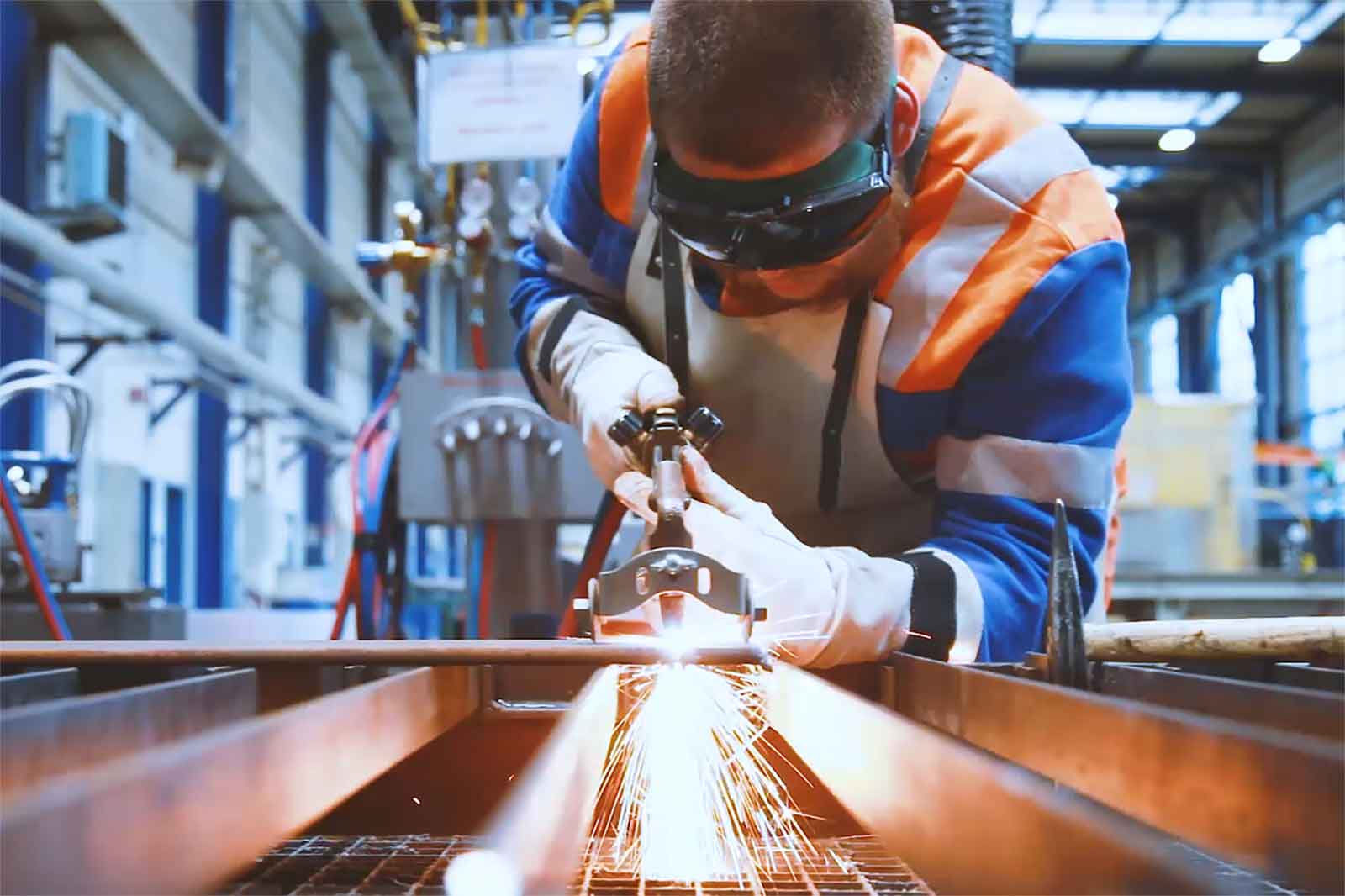 Ein Arbeiter in einer blauen und orangefarbenen Jacke nutzt eine Schweißpistole und erzeugt Funken in einer Werkstatt.