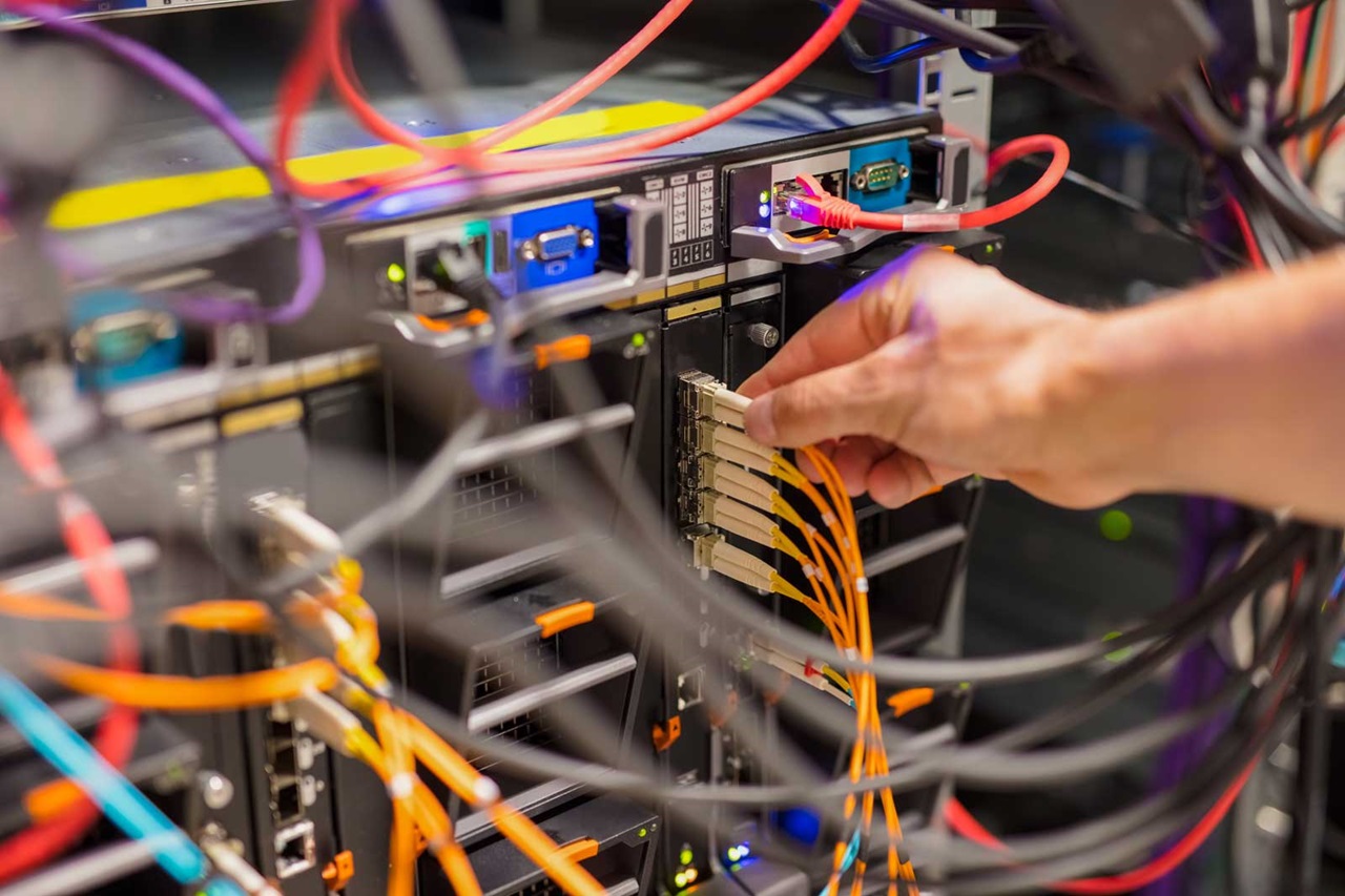 Eine Hand verbindet Glasfaserkabel mit einem Server in einem Netzwerk-Schrank, der mit bunten Kabeln und LED-Anzeigen gefüllt ist.
