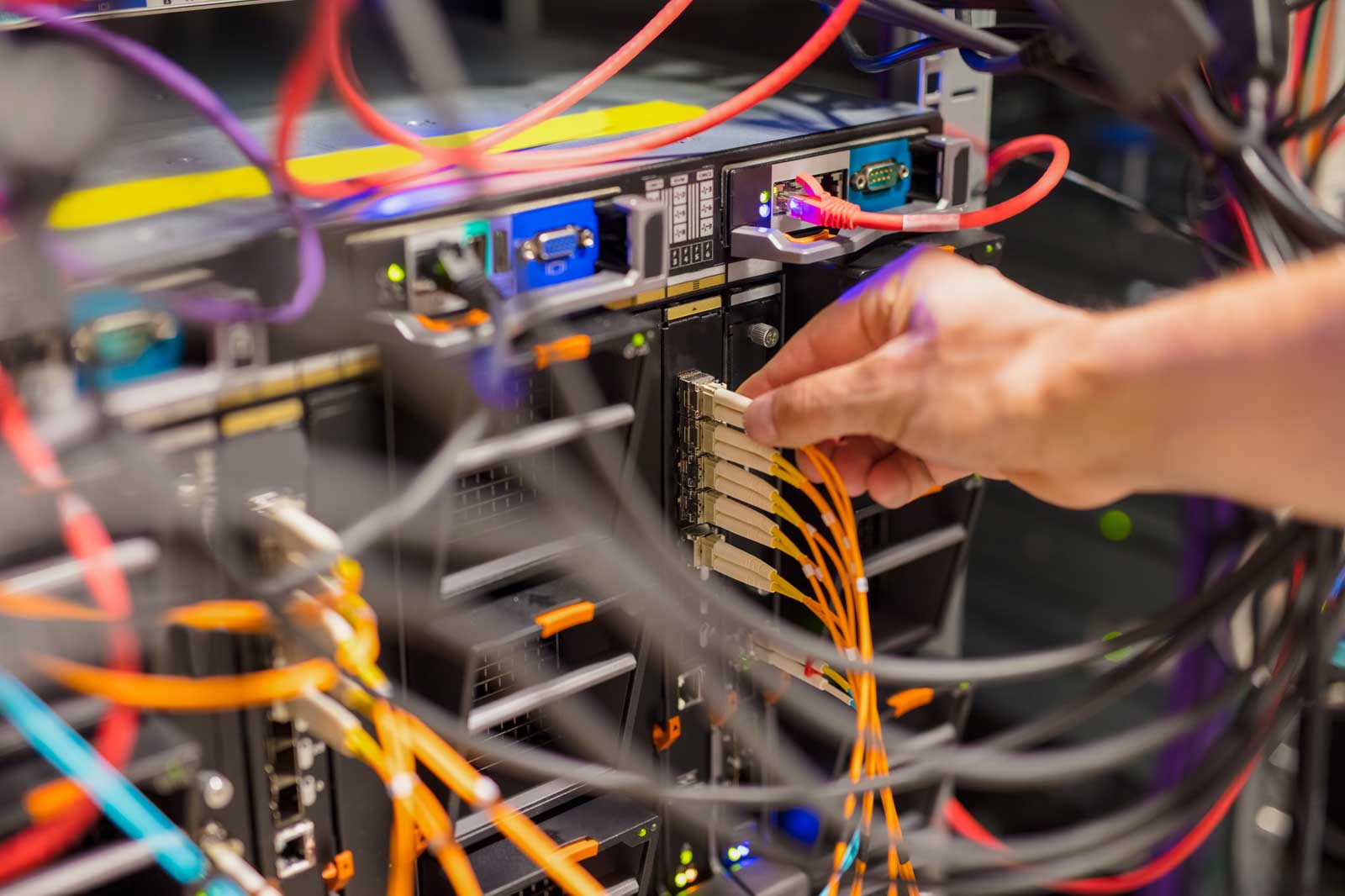 Eine Hand verbindet Glasfaserkabel mit einem Server in einem Netzwerk-Schrank, der mit bunten Kabeln und LED-Anzeigen gefüllt ist.