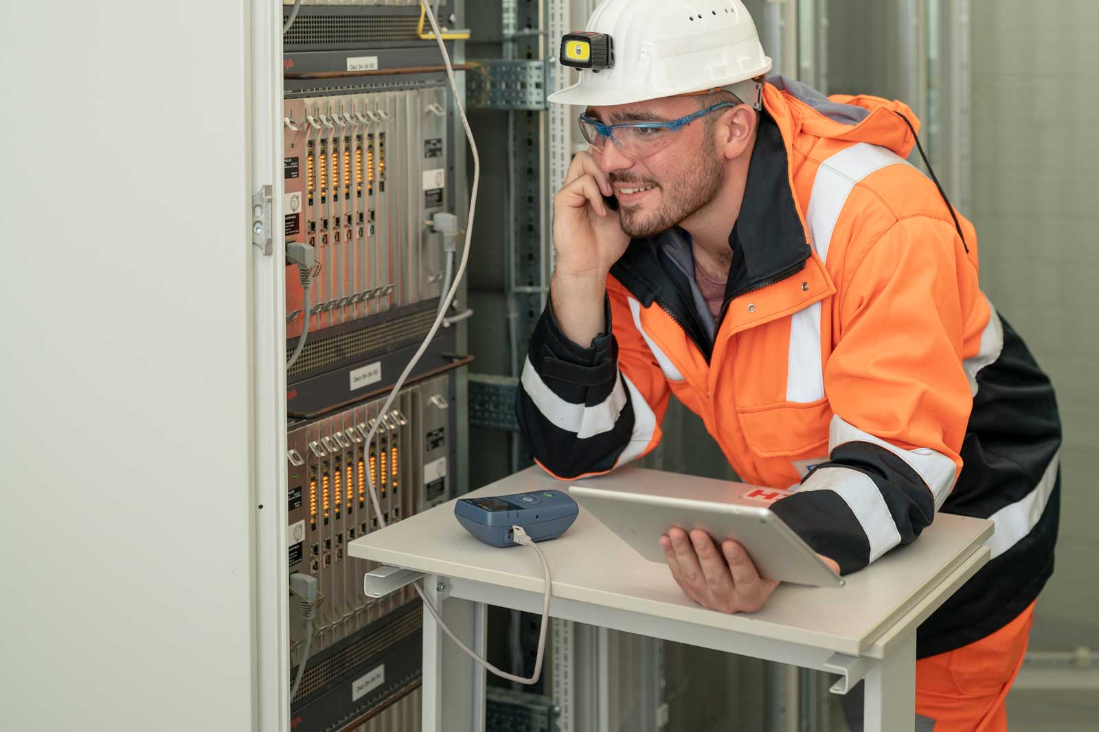 Ein Techniker in einer orangefarbenen Sicherheitsjacke untersucht mit einem Tablet Ausrüstung neben einem Serverrack mit leuchtenden Anzeigen.