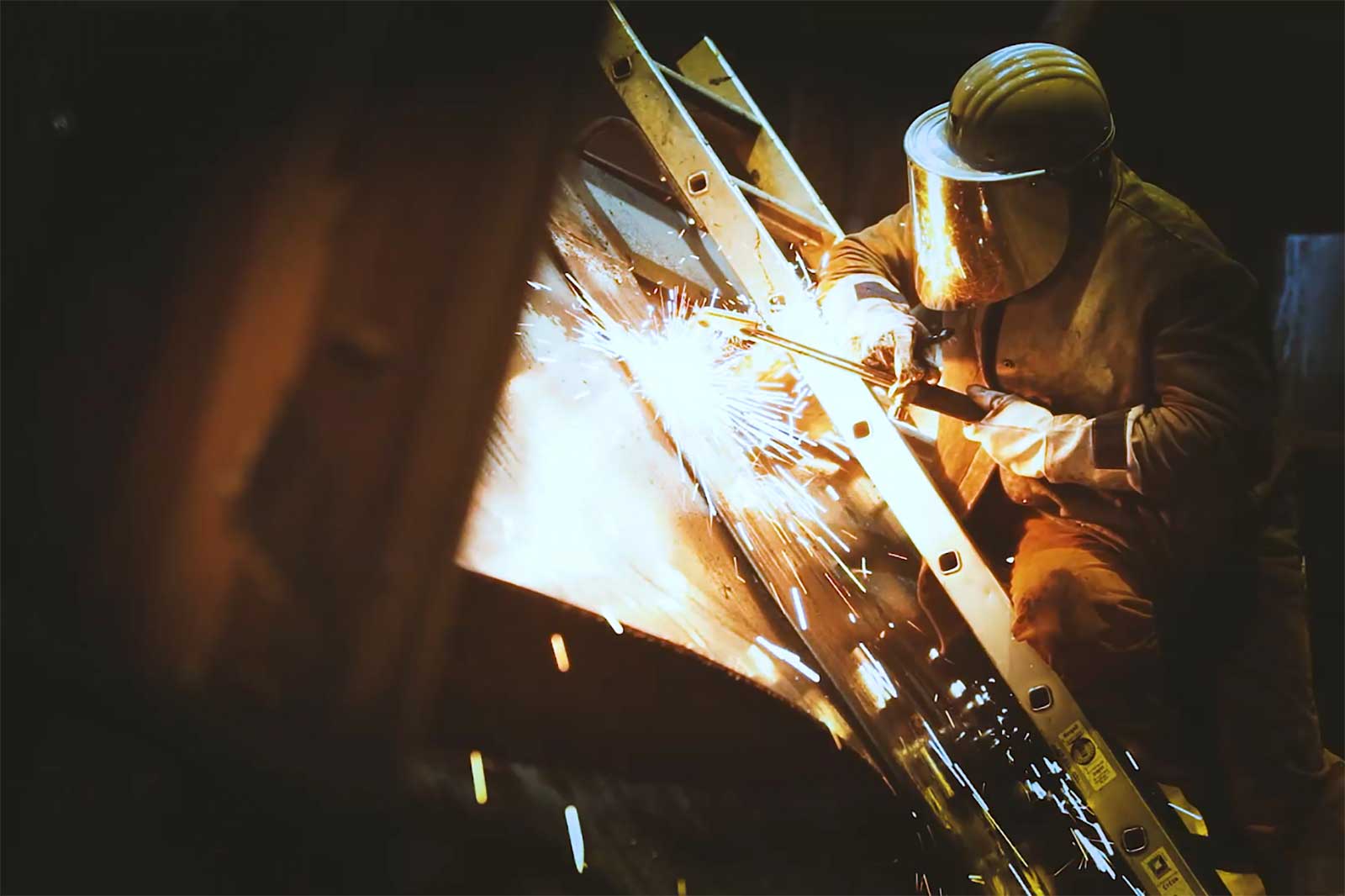 Ein Schweißer in Schutzkleidung arbeitet an Metall und erzeugt Funken in einer dunklen Werkstatt.